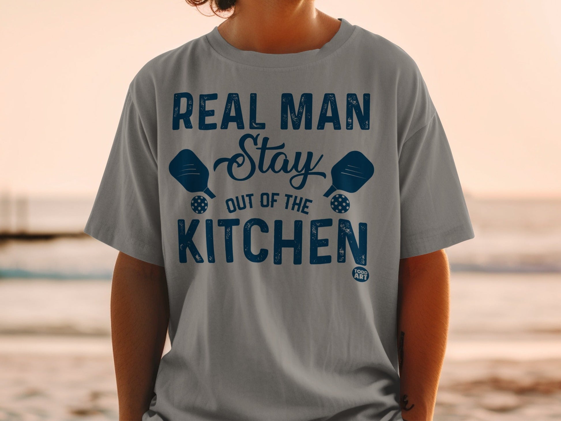 Grey t-shirt with blue text reading "REAL MAN STAY OUT OF THE KITCHEN" and pickleball paddles.