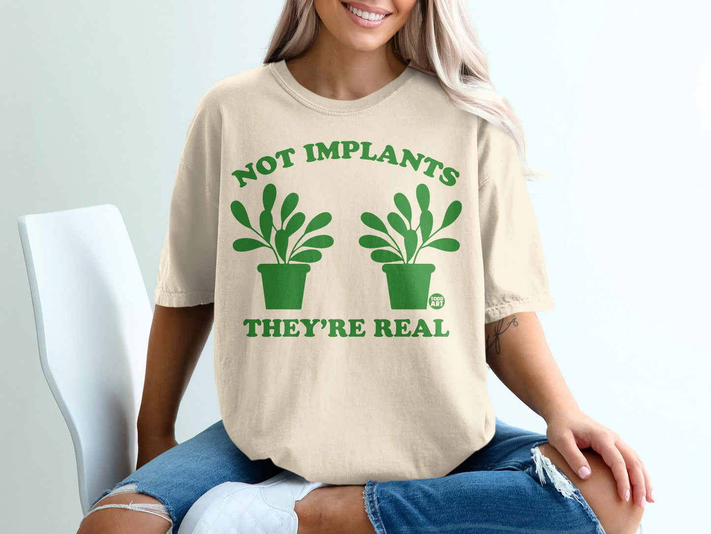 Cream t-shirt with green plants and text "NOT IMPLANTS THEY'RE REAL".