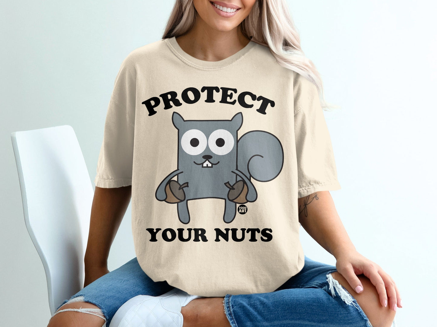 Beige t-shirt with cartoon squirrel holding acorns and text "PROTECT YOUR NUTS".