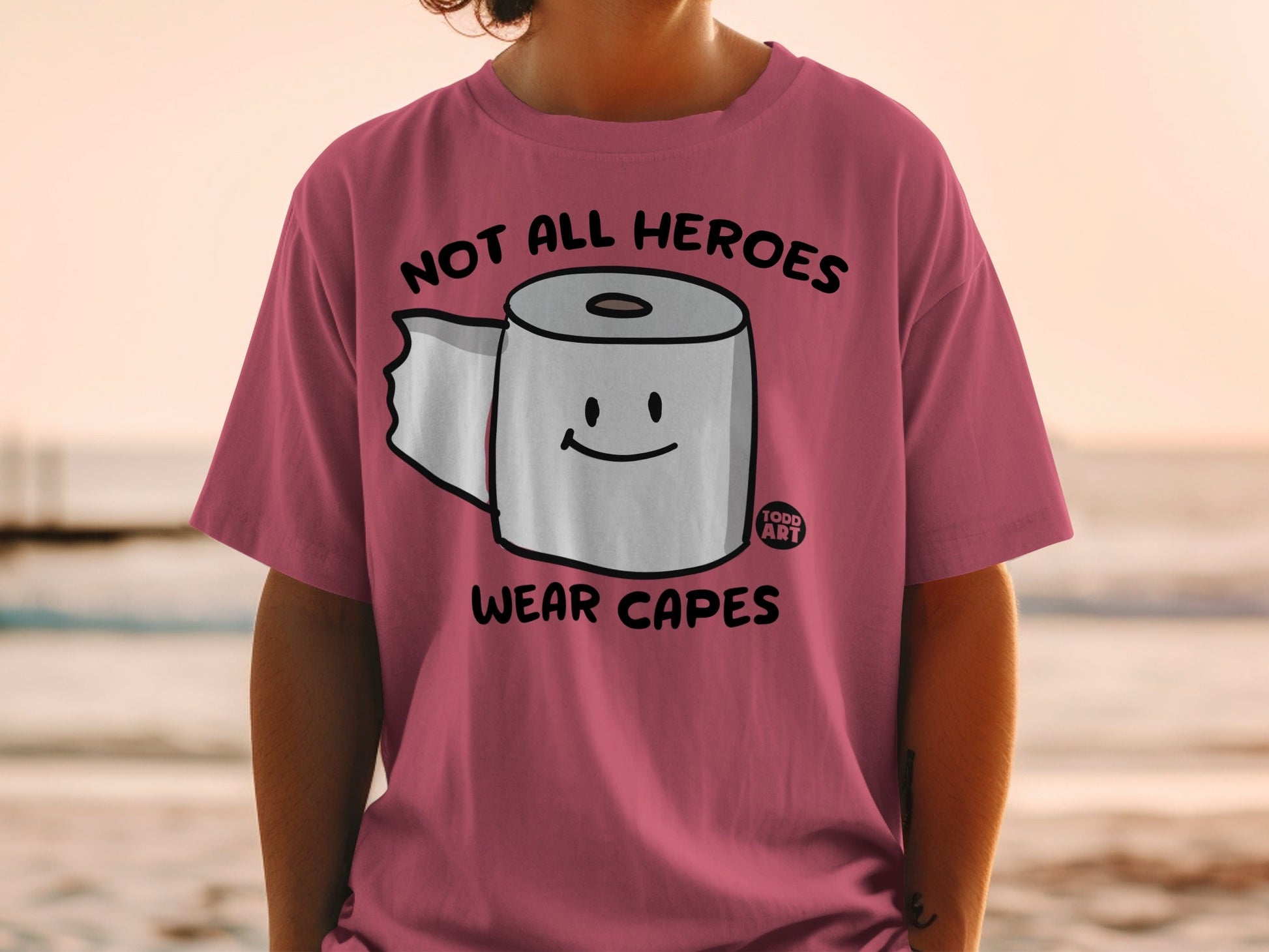 Pink t-shirt with toilet paper roll graphic and text "Not all heroes wear capes."