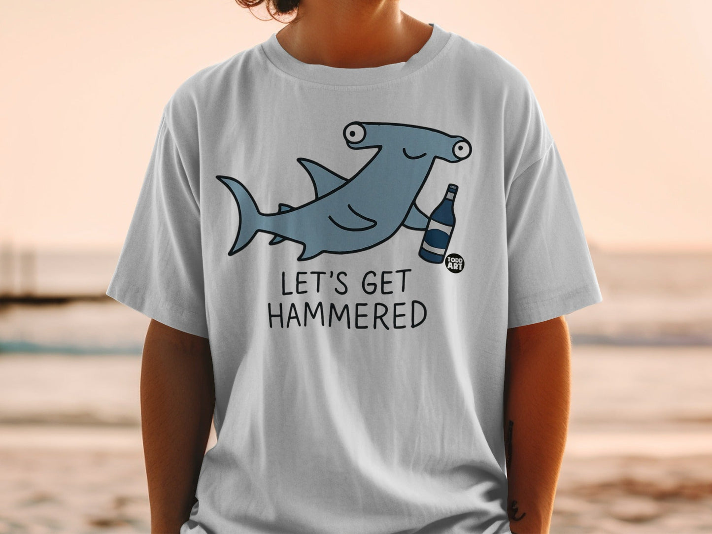 T-shirt graphic of a cartoon hammerhead shark holding a bottle, with text "LET'S GET HAMMERED".