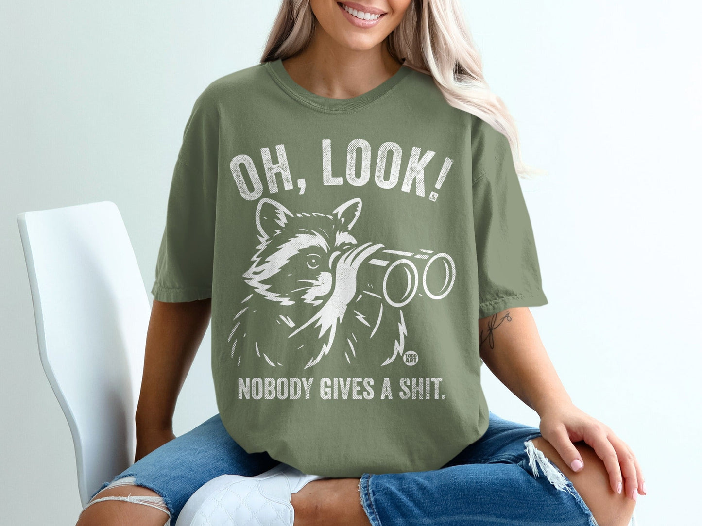 Olive green t-shirt with a raccoon looking through binoculars and text reading "OH, LOOK! NOBODY GIVES A SHIT."