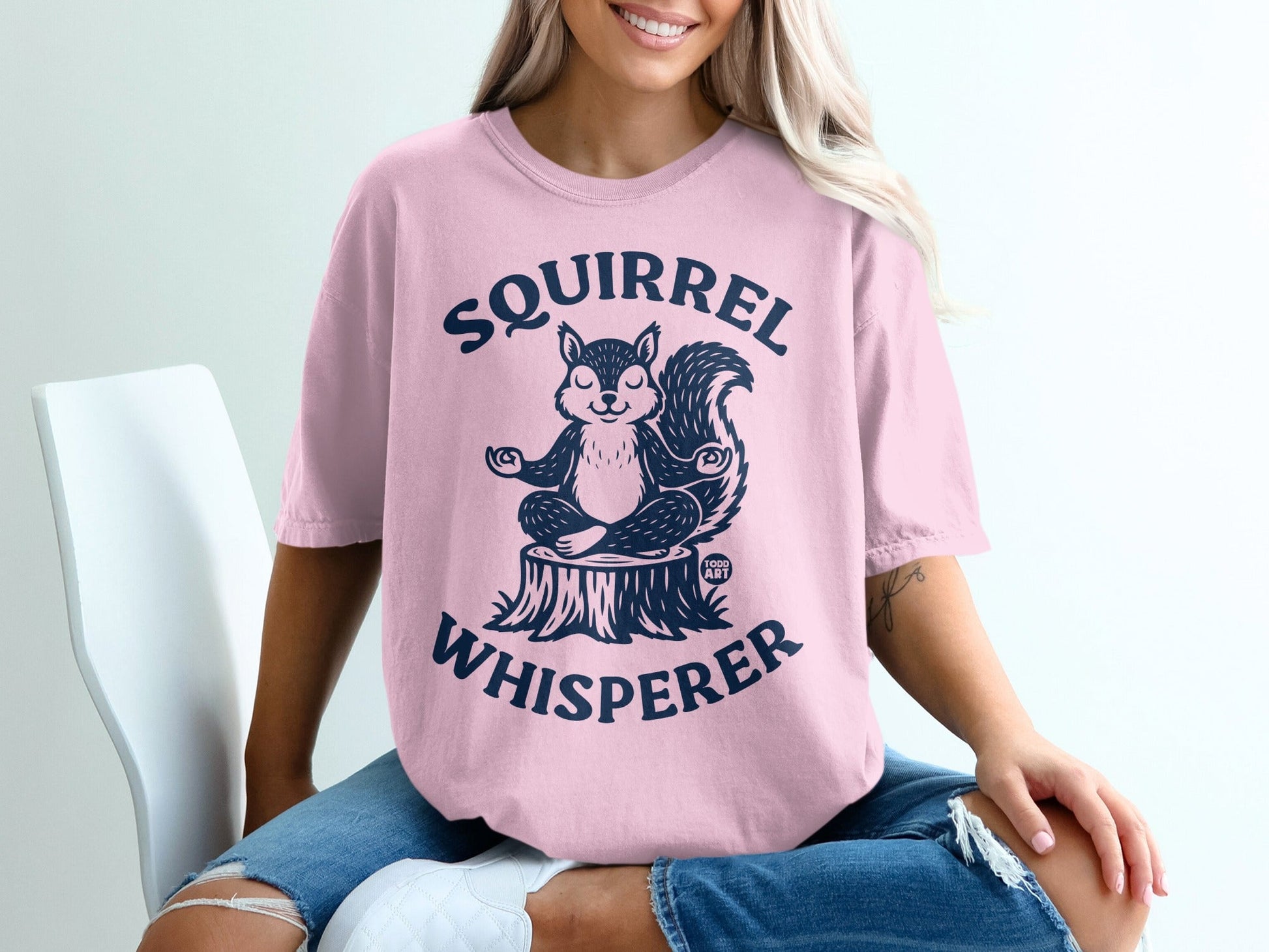 Pink t-shirt with a meditating squirrel on a tree stump, "SQUIRREL WHISPERER" text.