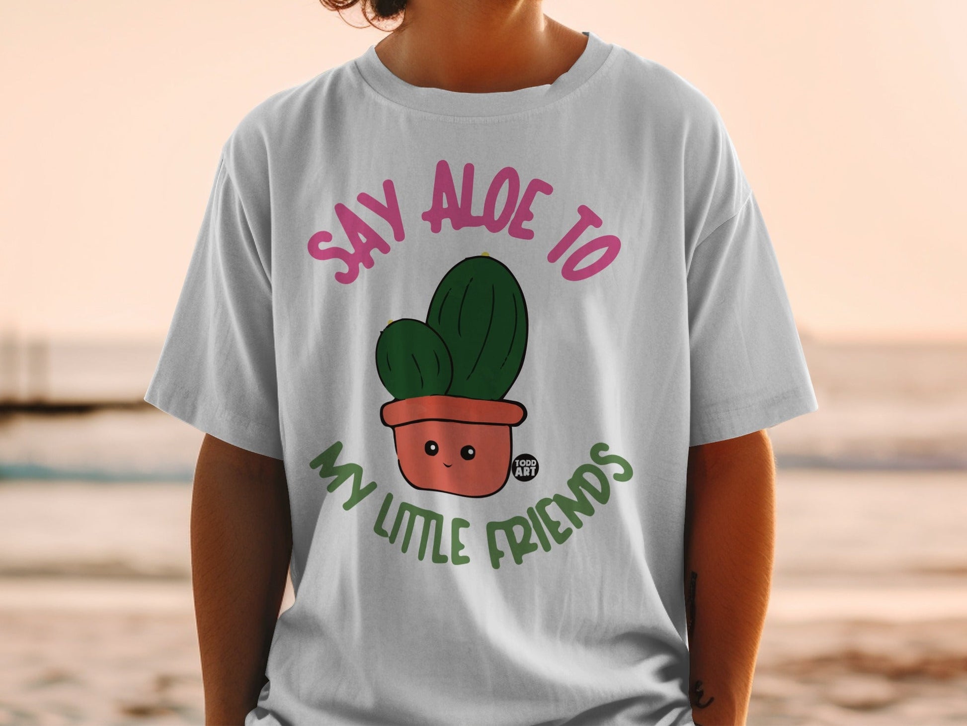 White t-shirt with pink text saying "Say Aloe To My Little Friends" and a potted cactus.