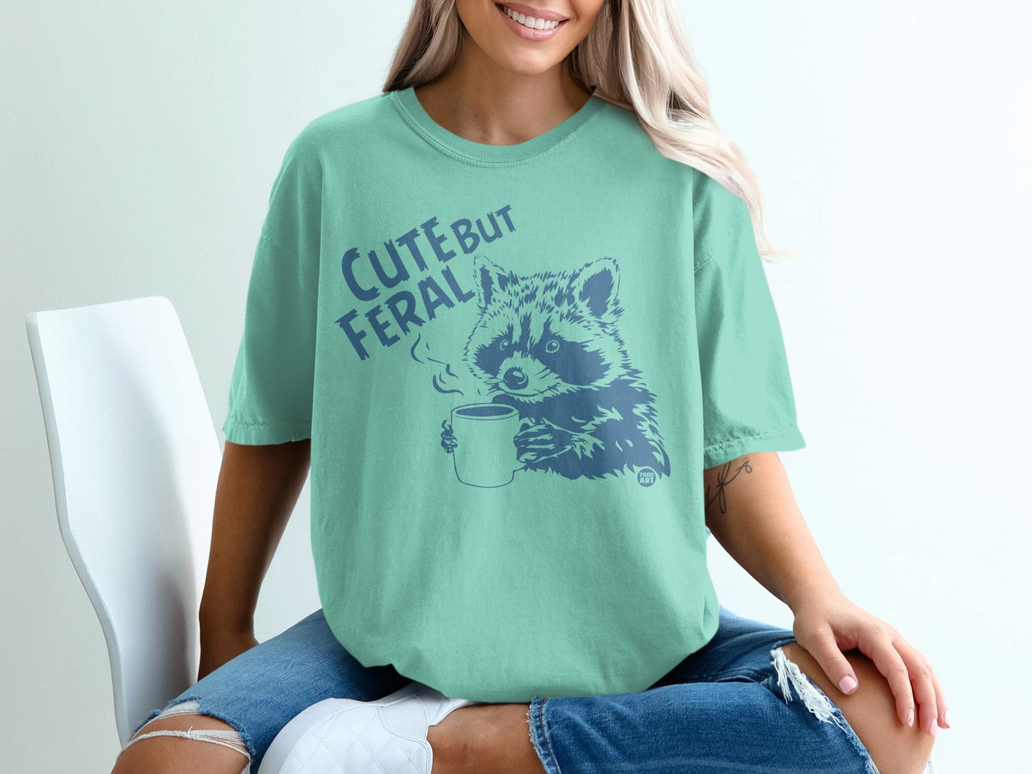 Teal t-shirt with a raccoon drinking coffee and the text "Cute But Feral."