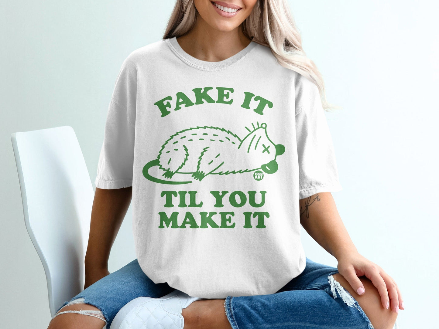 White t-shirt with green graphic of a possum and text "Fake It Til You Make It".