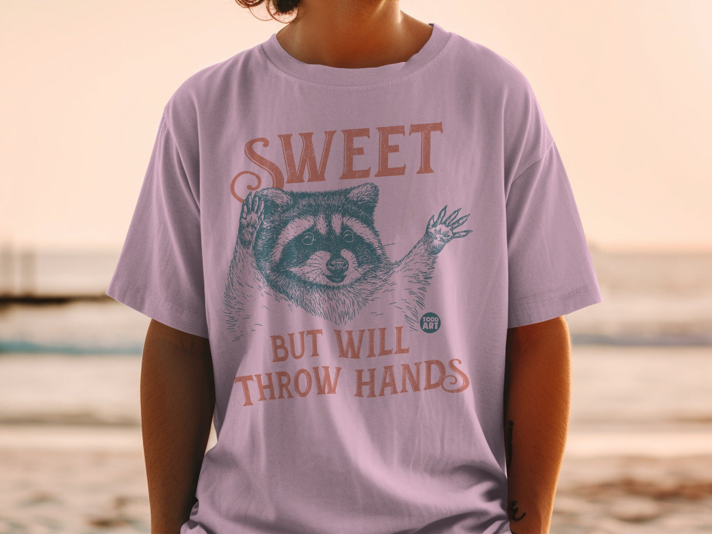 T-shirt with raccoon graphic and text "SWEET BUT WILL THROW HANDS".
