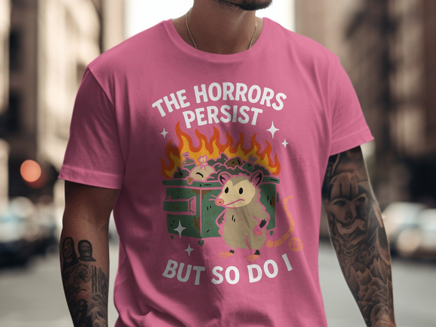 Pink t-shirt with possum and dumpster fire graphic, text reads "The horrors persist but so do I".