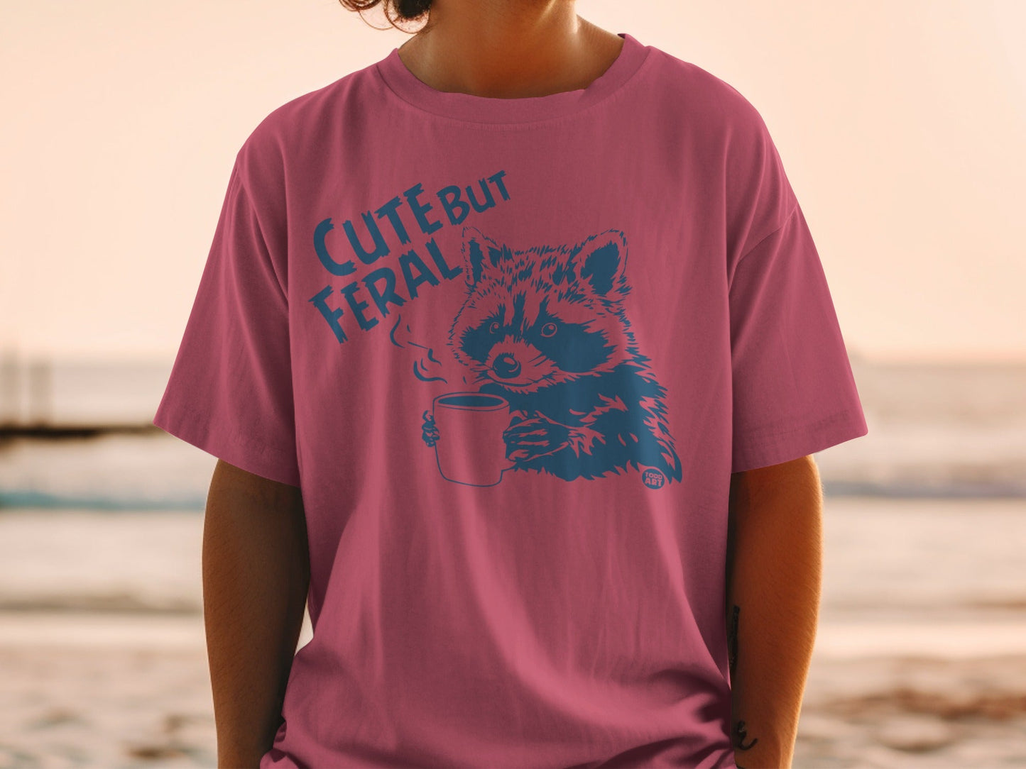 T-shirt with raccoon holding coffee mug, text "Cute but Feral".