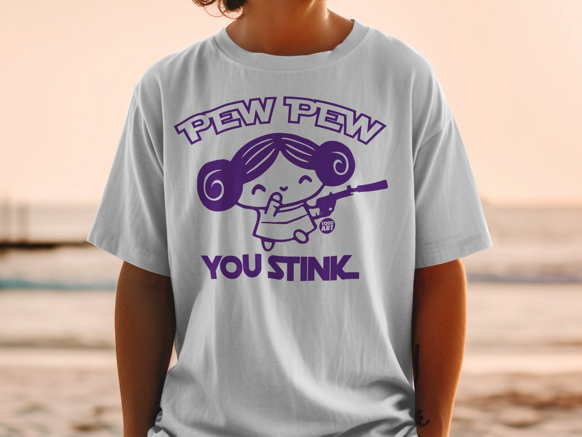 White t-shirt with purple cartoon girl holding a gun, text "PEW PEW YOU STINK".