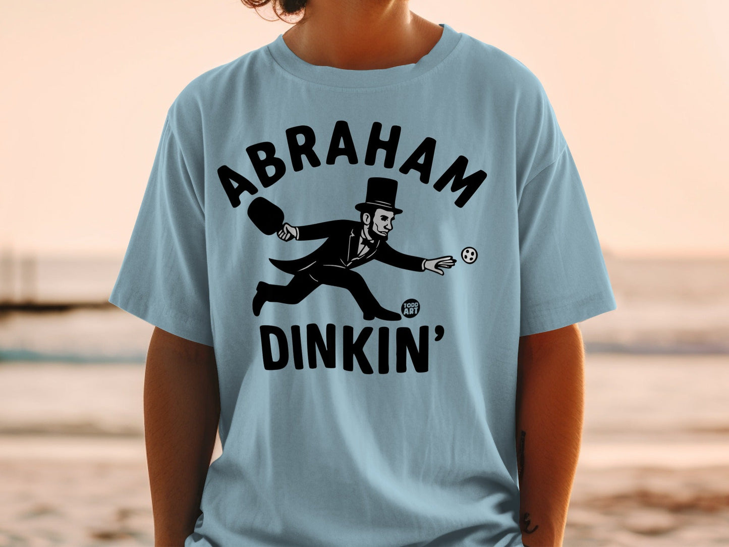 T-shirt with Abraham Lincoln playing pickleball, text "ABRAHAM DINKIN'".