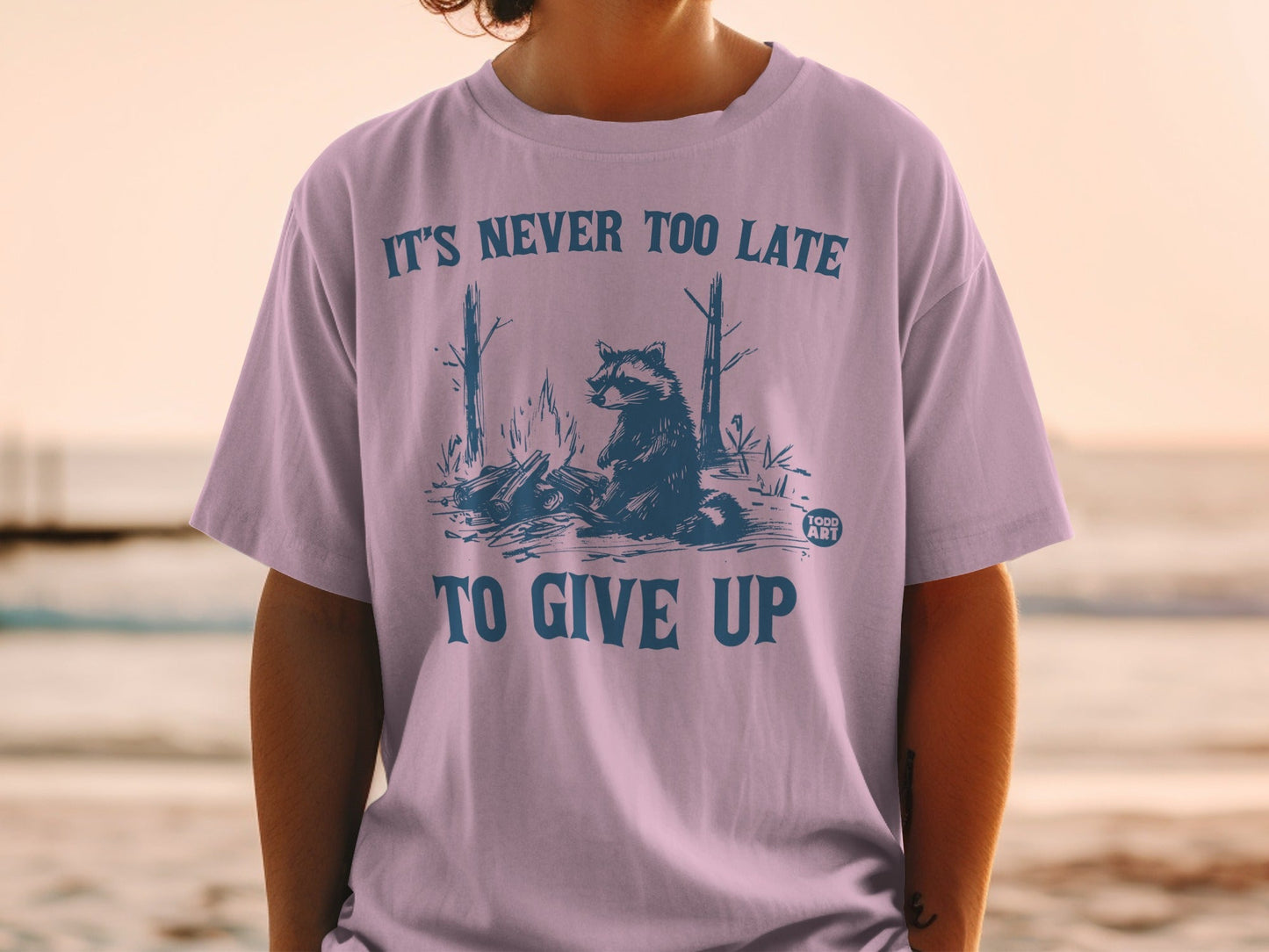 T-shirt with raccoon by campfire graphic and "IT'S NEVER TOO LATE TO GIVE UP" text.