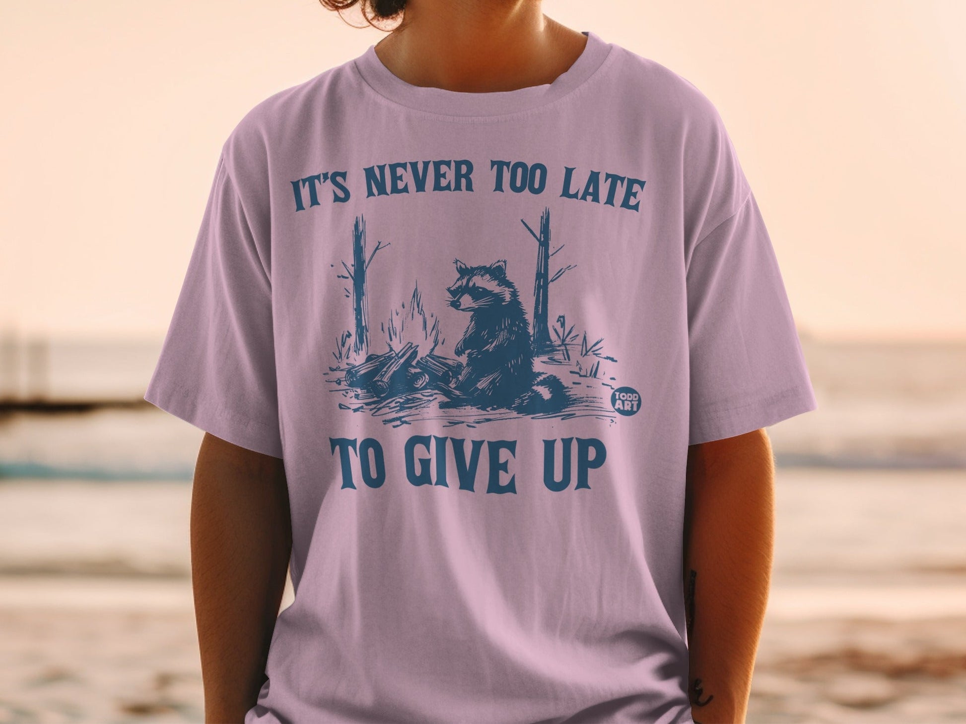 T-shirt with raccoon by campfire graphic and "IT'S NEVER TOO LATE TO GIVE UP" text.