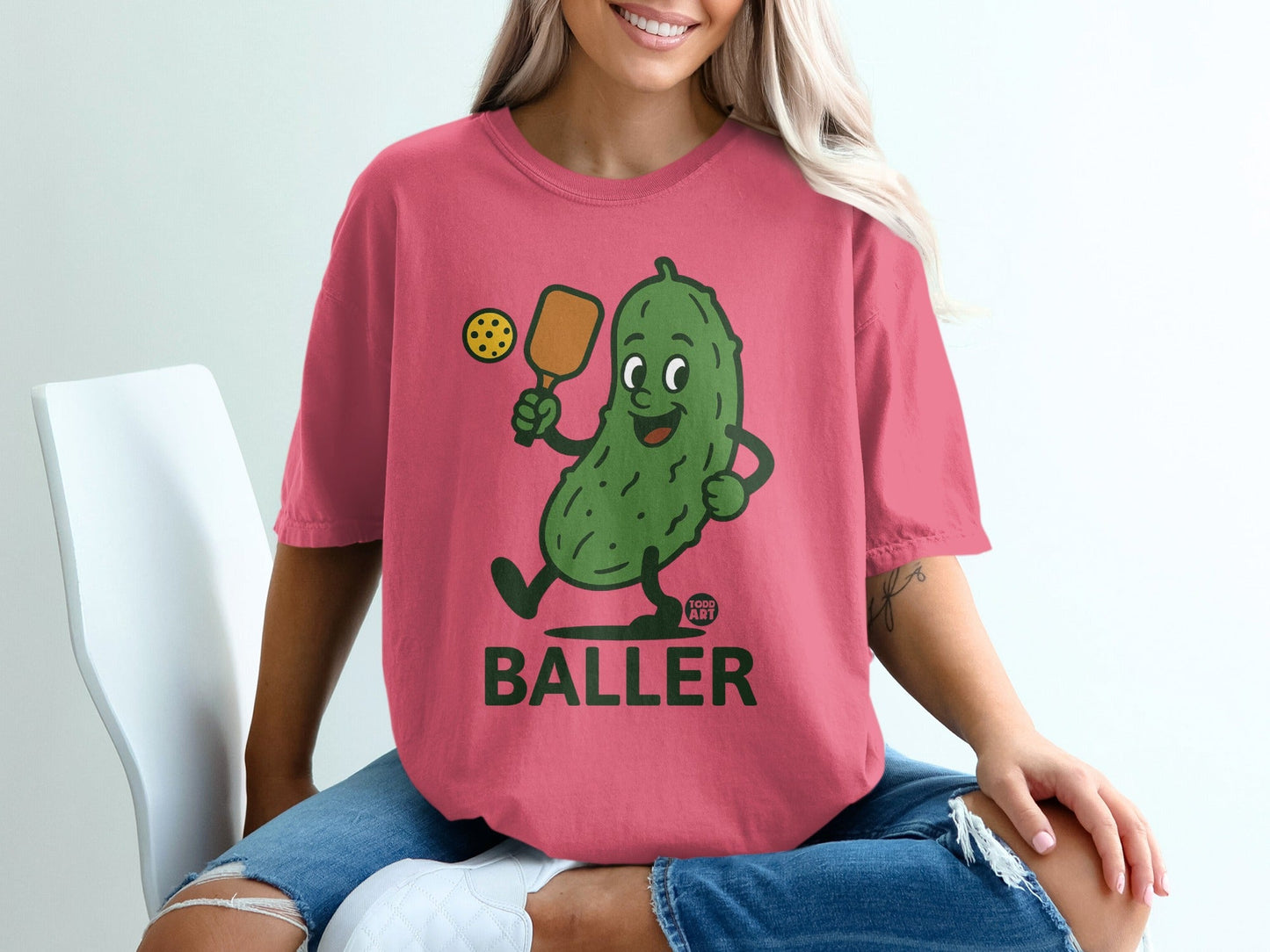 Cartoon pickle with paddle and ball, wearing a pink t-shirt.