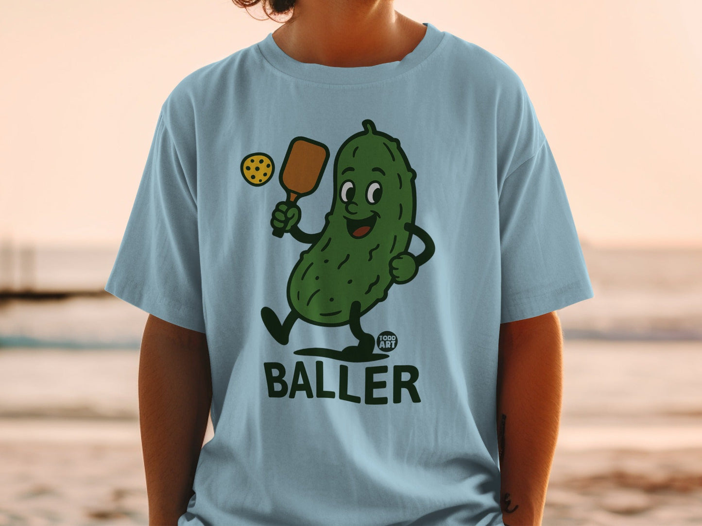 Cartoon pickle playing pickleball on a light blue t-shirt.