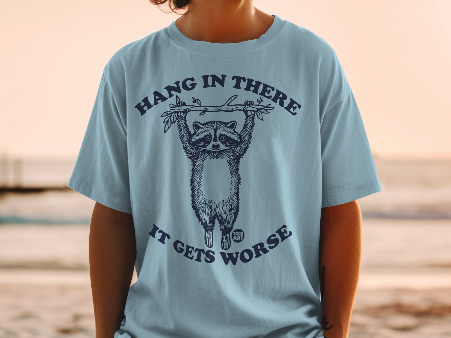 T-shirt with raccoon hanging from branch, text "Hang in there it gets worse".