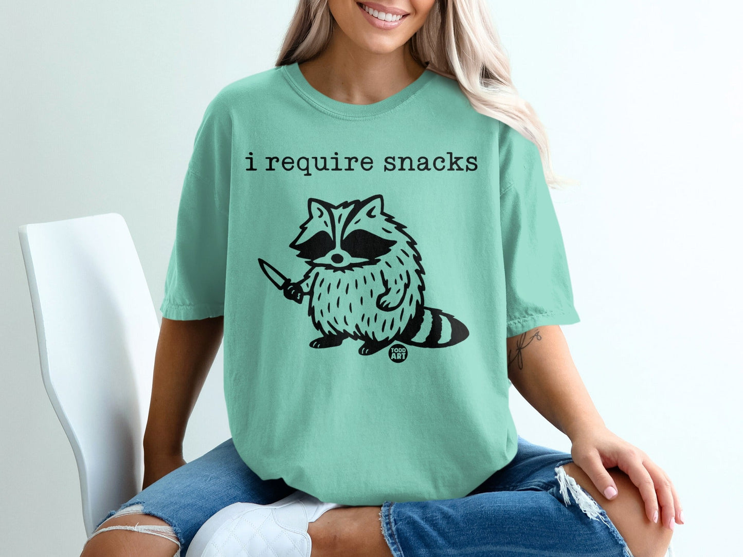 Teal t-shirt with a cartoon raccoon holding a knife and the text "i require snacks".