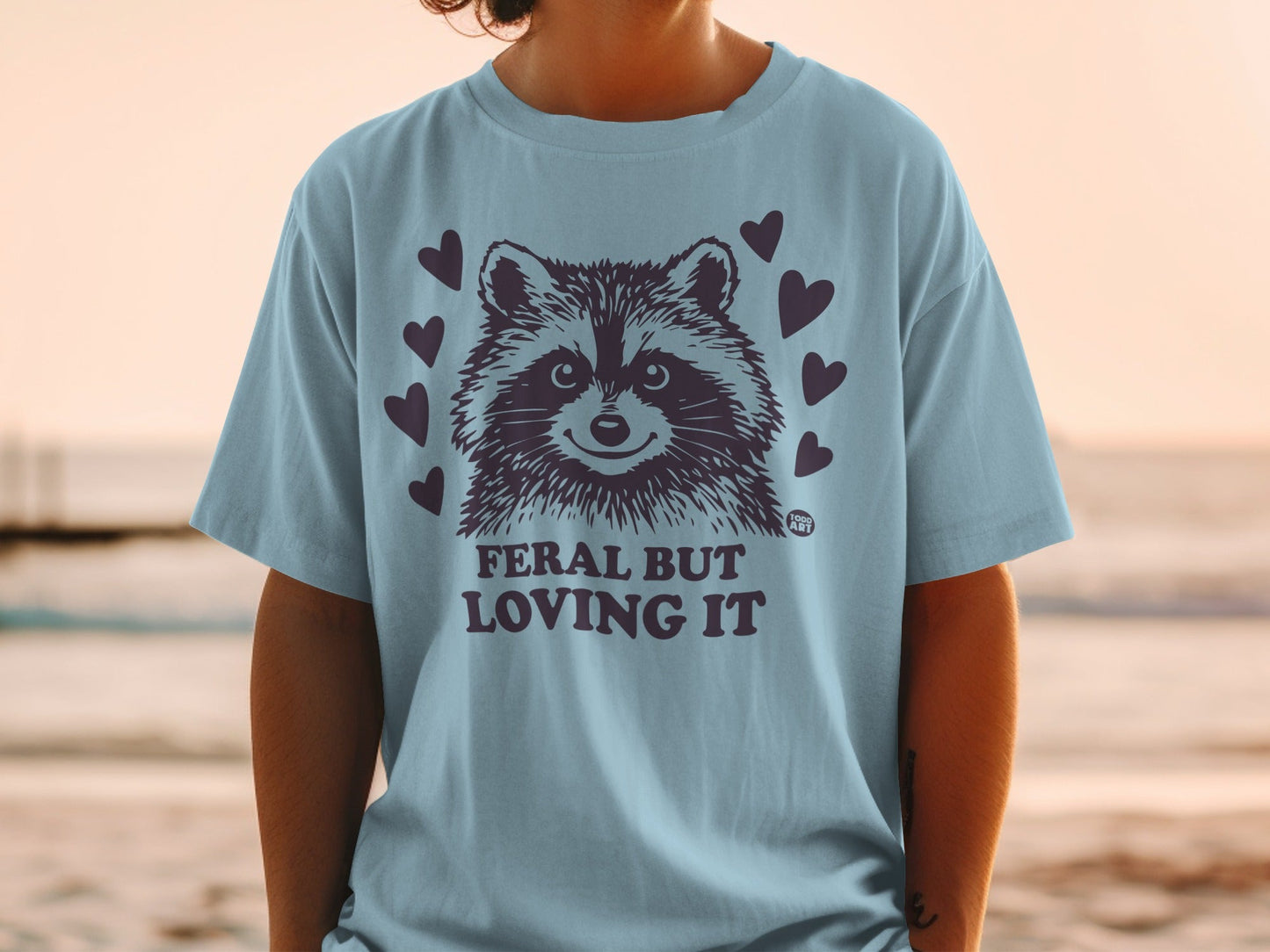Light blue t-shirt with raccoon graphic and "FERAL BUT LOVING IT" text.
