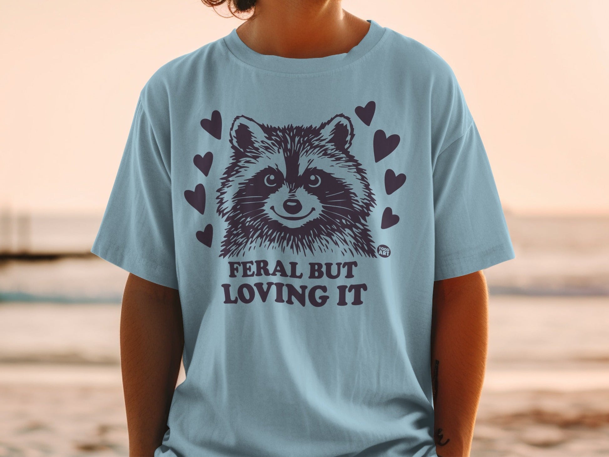 Light blue t-shirt with raccoon graphic and "FERAL BUT LOVING IT" text.