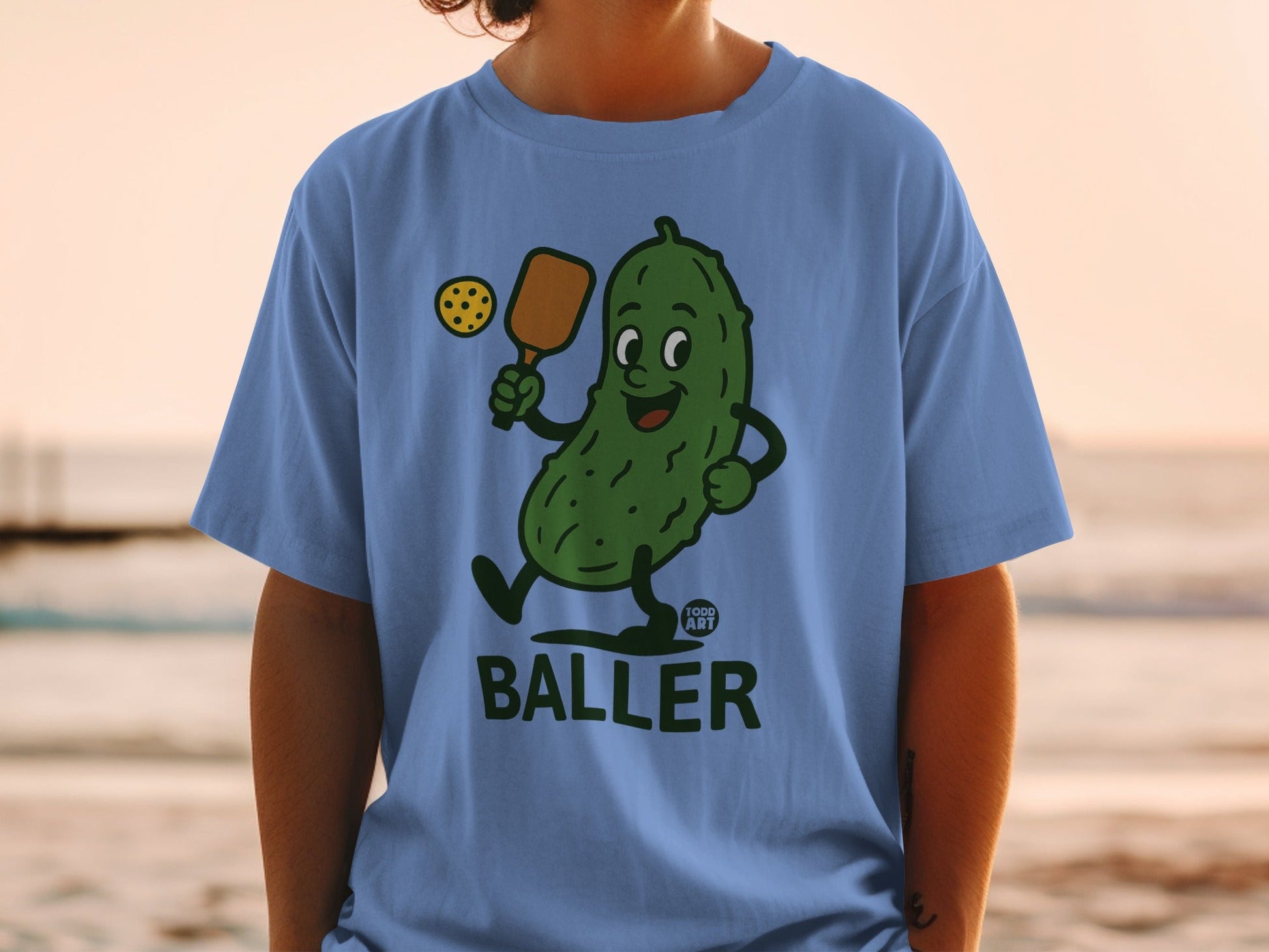 Blue t-shirt with cartoon pickle playing pickleball.