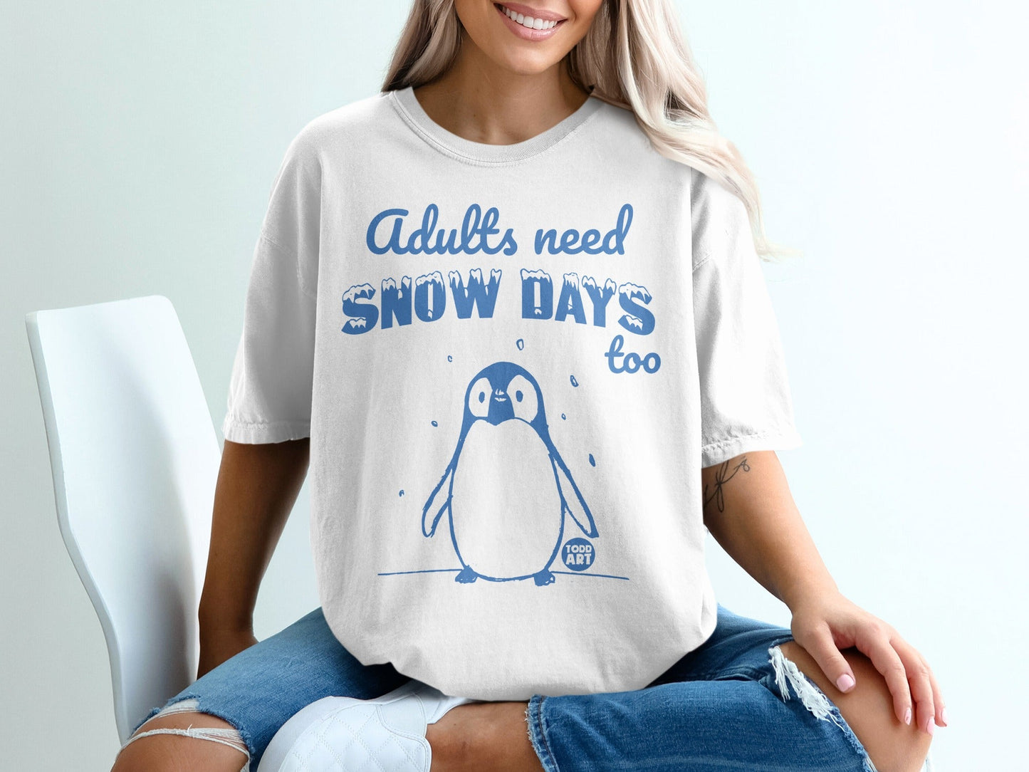White t-shirt with blue text "Adults need SNOW DAYS too" and a cartoon penguin.