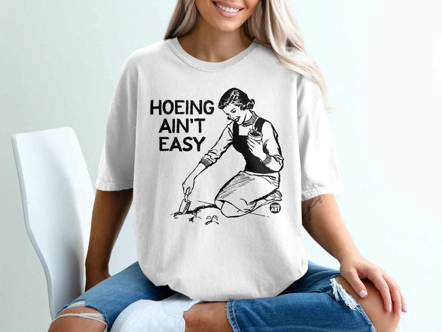 White t-shirt with graphic of a woman gardening and text "HOEING AIN'T EASY".