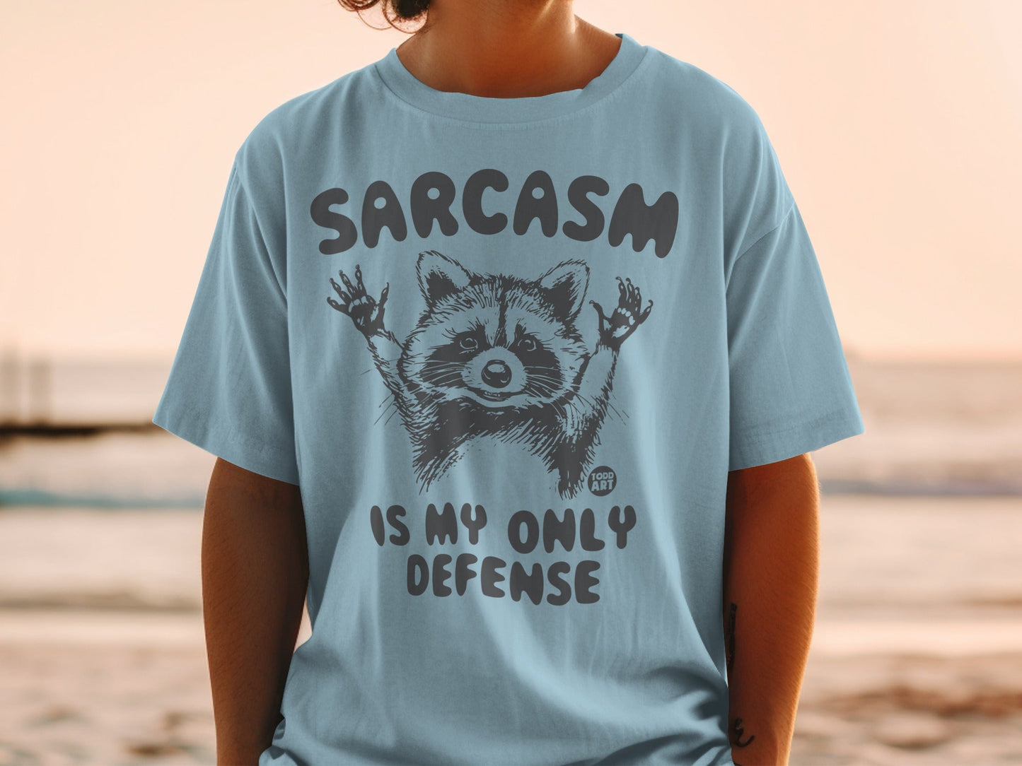 Light blue t-shirt with raccoon graphic and text "SARCASM IS MY ONLY DEFENSE".