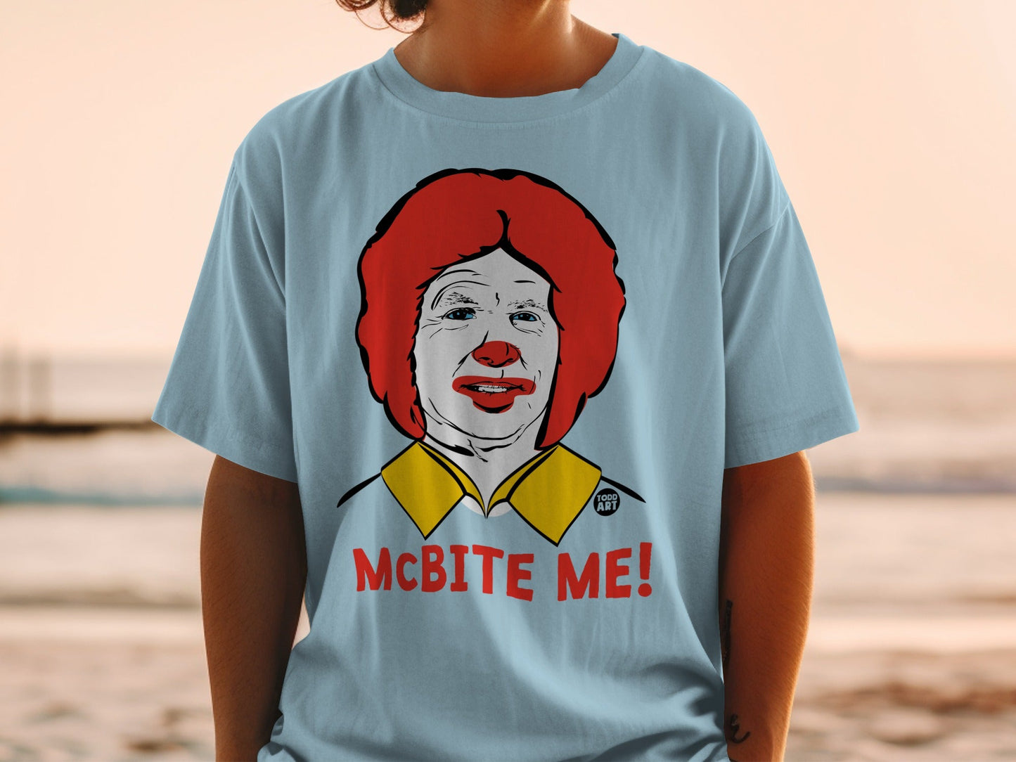 Light blue t-shirt featuring a close-up illustration of Ronald McDonald's face with the text "MCBITE ME!"