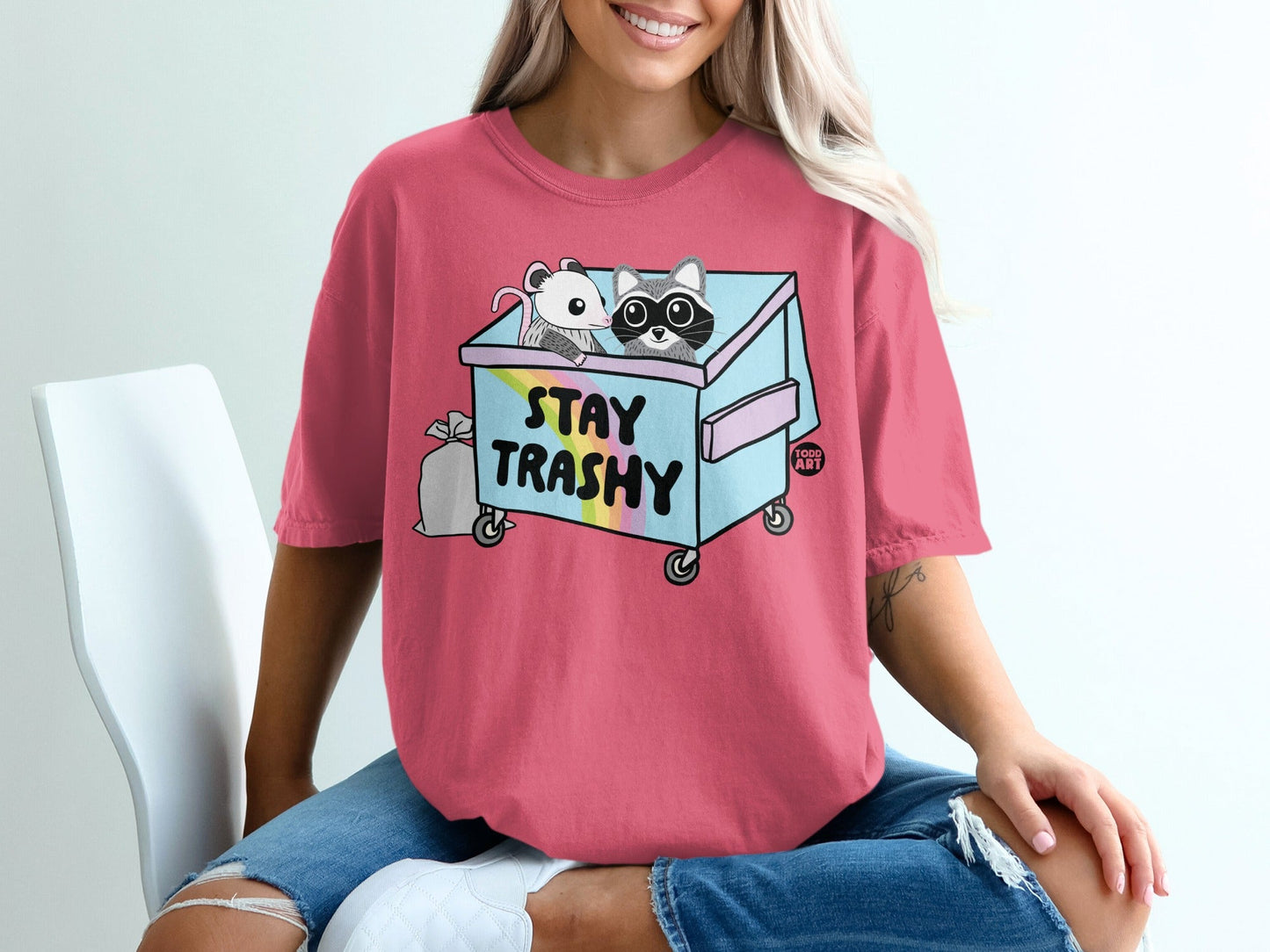 Pink t-shirt with "Stay Trashy" text and cartoon raccoons in a dumpster.