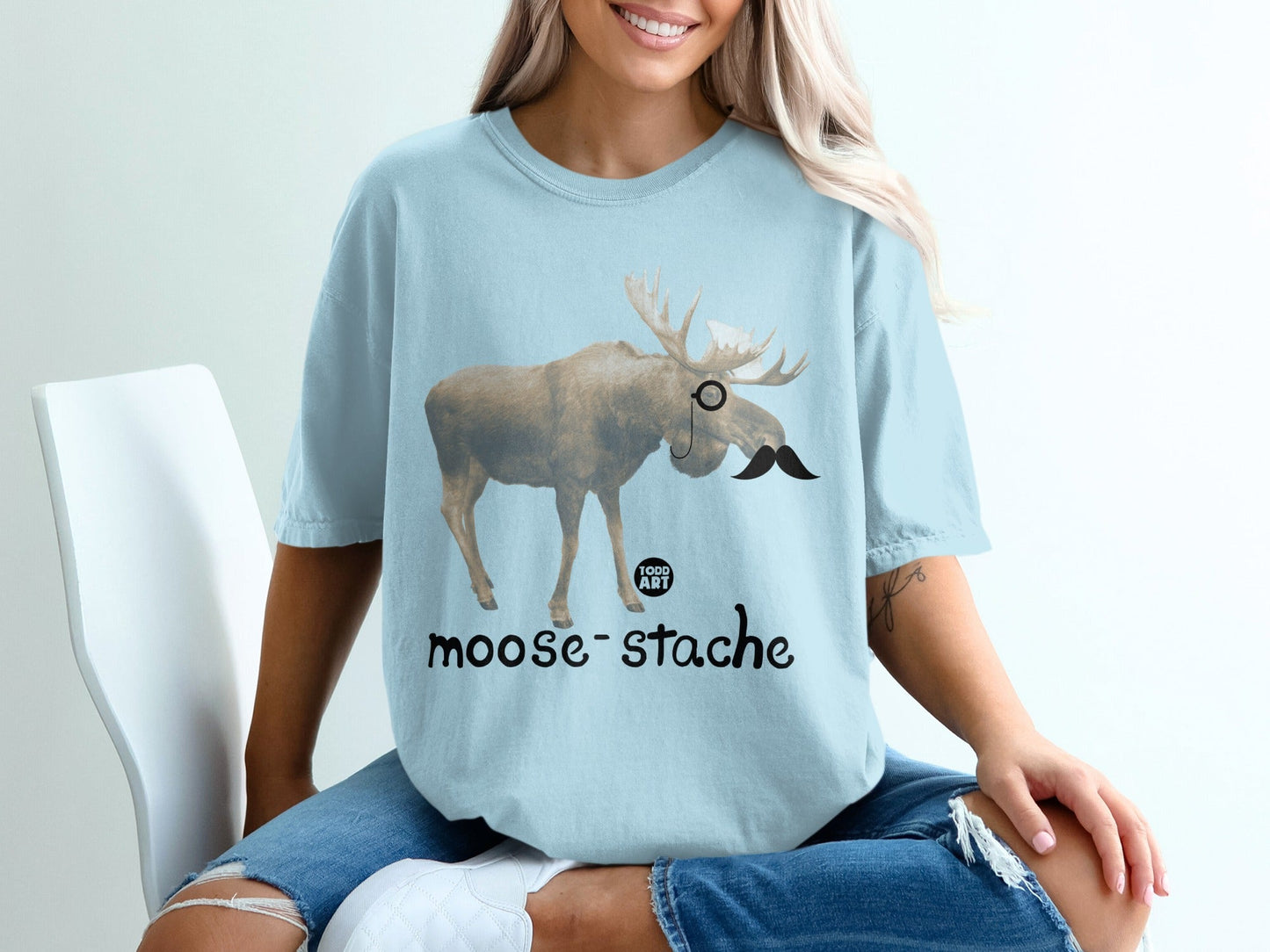 Light blue t-shirt with a moose wearing a monocle and mustache, text "moose-stache".