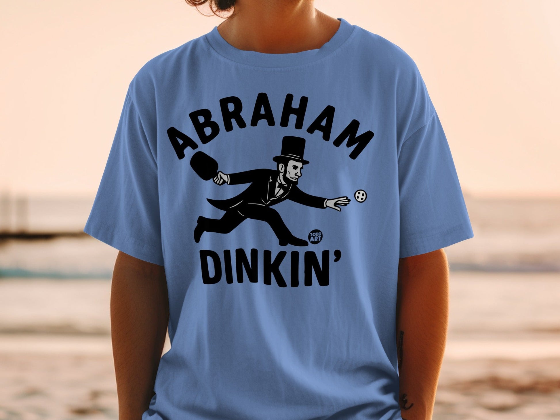 Blue t-shirt with Abraham Lincoln playing pickleball graphic and "Abraham Dinkin'" text.
