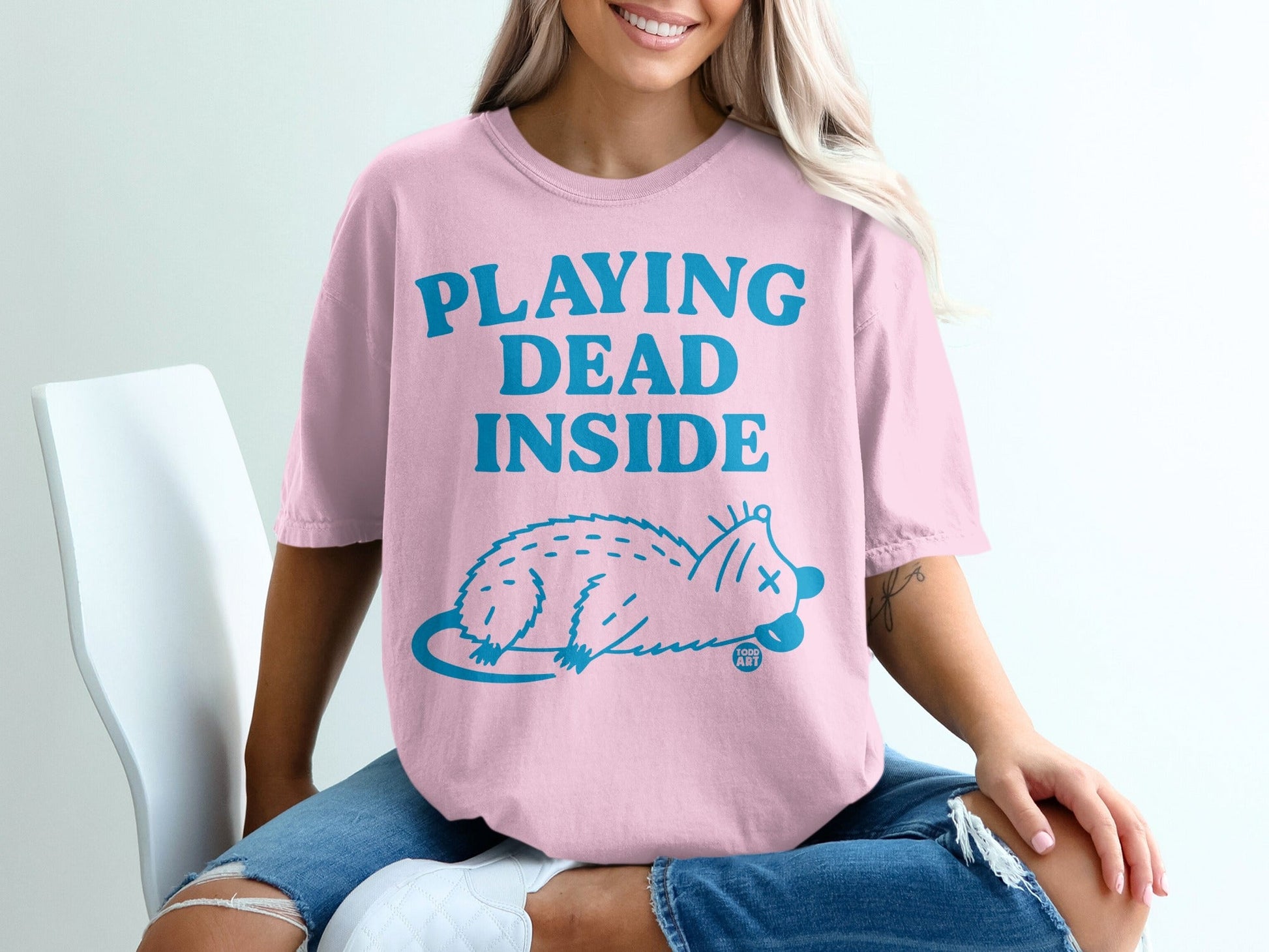 Pink t-shirt with blue text "PLAYING DEAD INSIDE" and a cartoon opossum graphic.