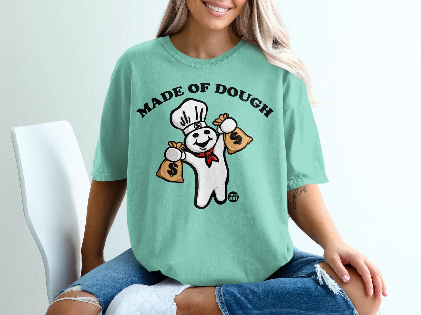 Pillsbury Doughboy with chef hat holding money bags on mint t-shirt.