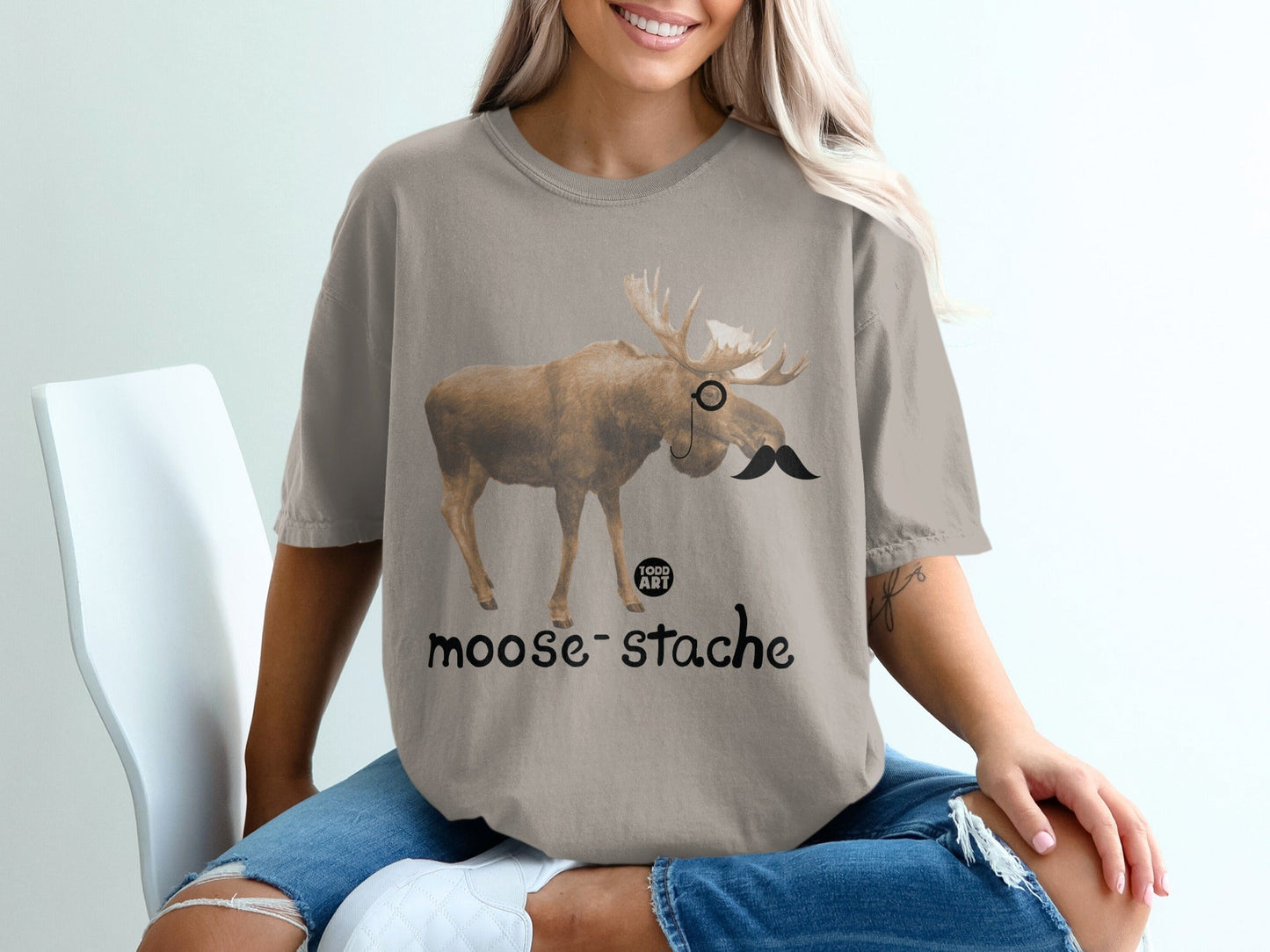 Moose with a mustache and monocle on a taupe t-shirt.