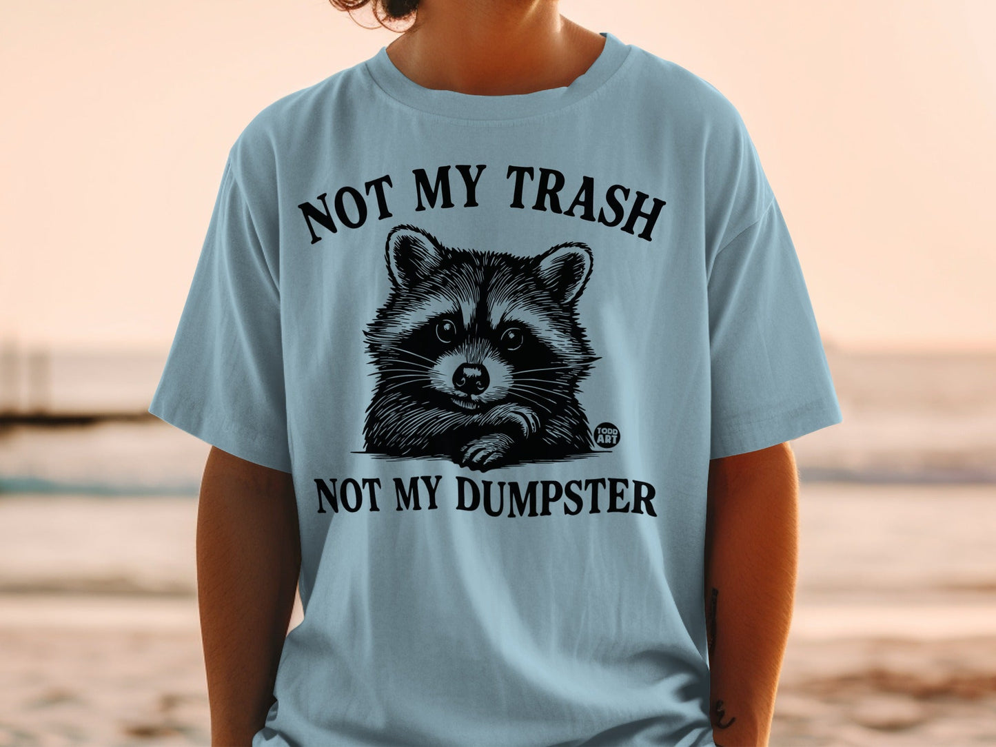 Light blue t-shirt with a raccoon graphic and text "NOT MY TRASH NOT MY DUMPSTER".