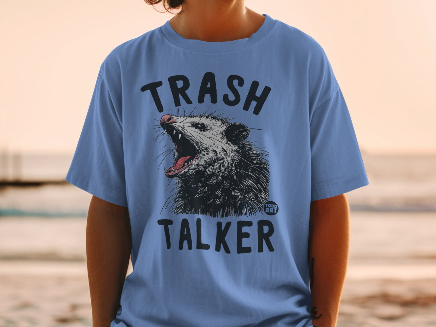 Blue t-shirt with a graphic of an opossum yelling and the words "TRASH TALKER".