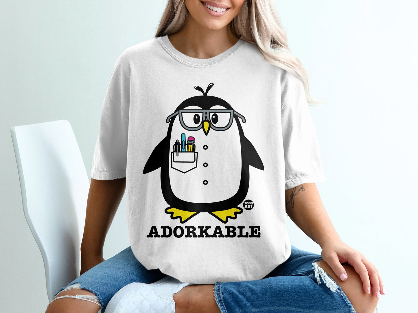 White t-shirt with a graphic of a penguin wearing glasses and holding pens.