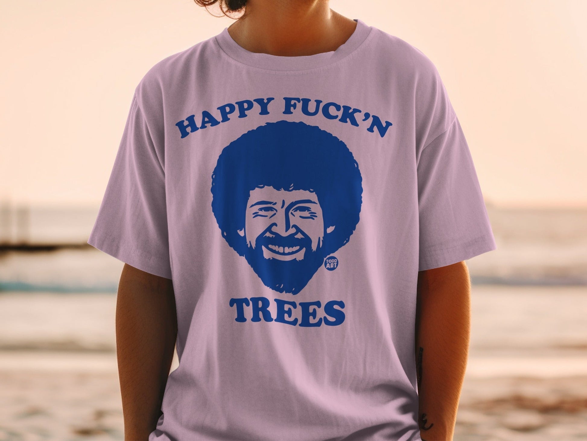 Lavender t-shirt with Bob Ross illustration and "HAPPY FUCK'N TREES" text.