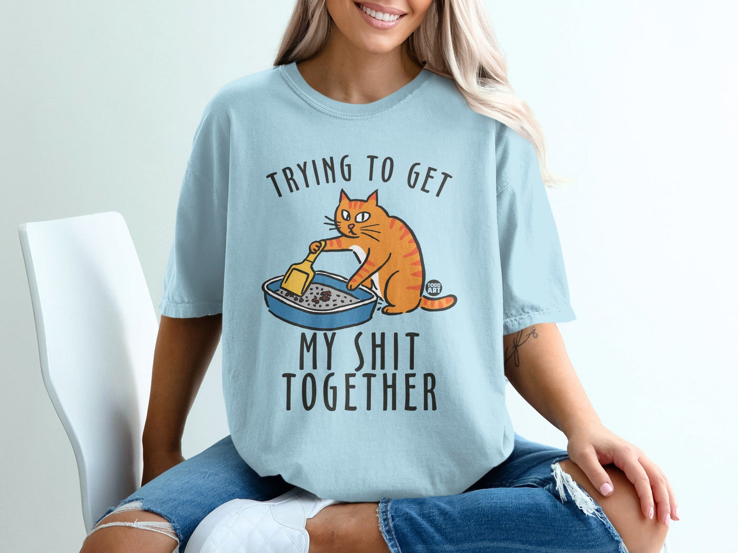 Light blue t-shirt with an orange cat cleaning a litter box and the text "TRYING TO GET MY SHIT TOGETHER".