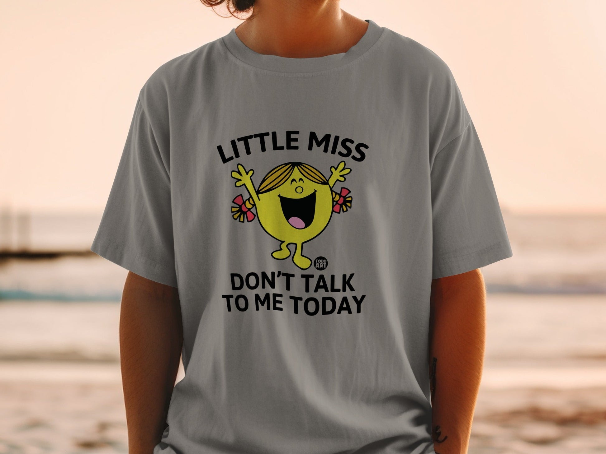 Little Miss character in yellow, smiling, on a gray t-shirt with text.