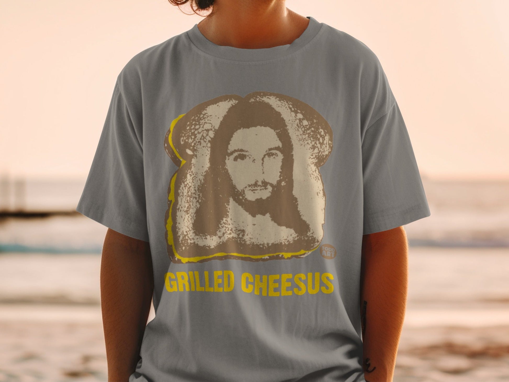 Grilled Cheesus graphic on a gray t-shirt with a beach background.