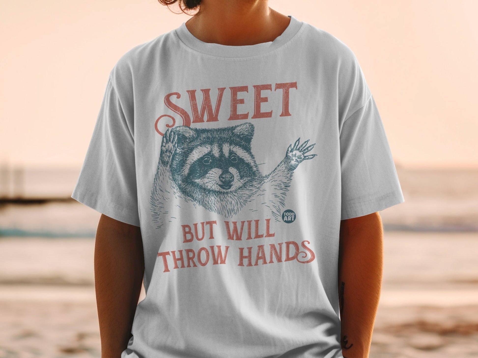 T-shirt with raccoon graphic and text "SWEET BUT WILL THROW HANDS".
