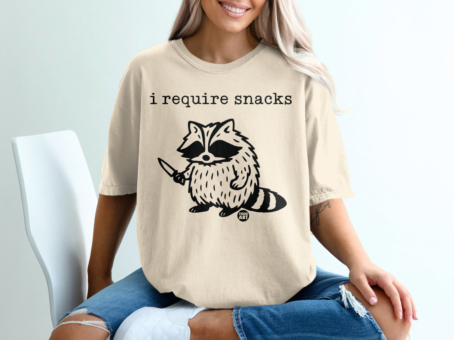 Cream t-shirt with raccoon holding knife and text "I require snacks".