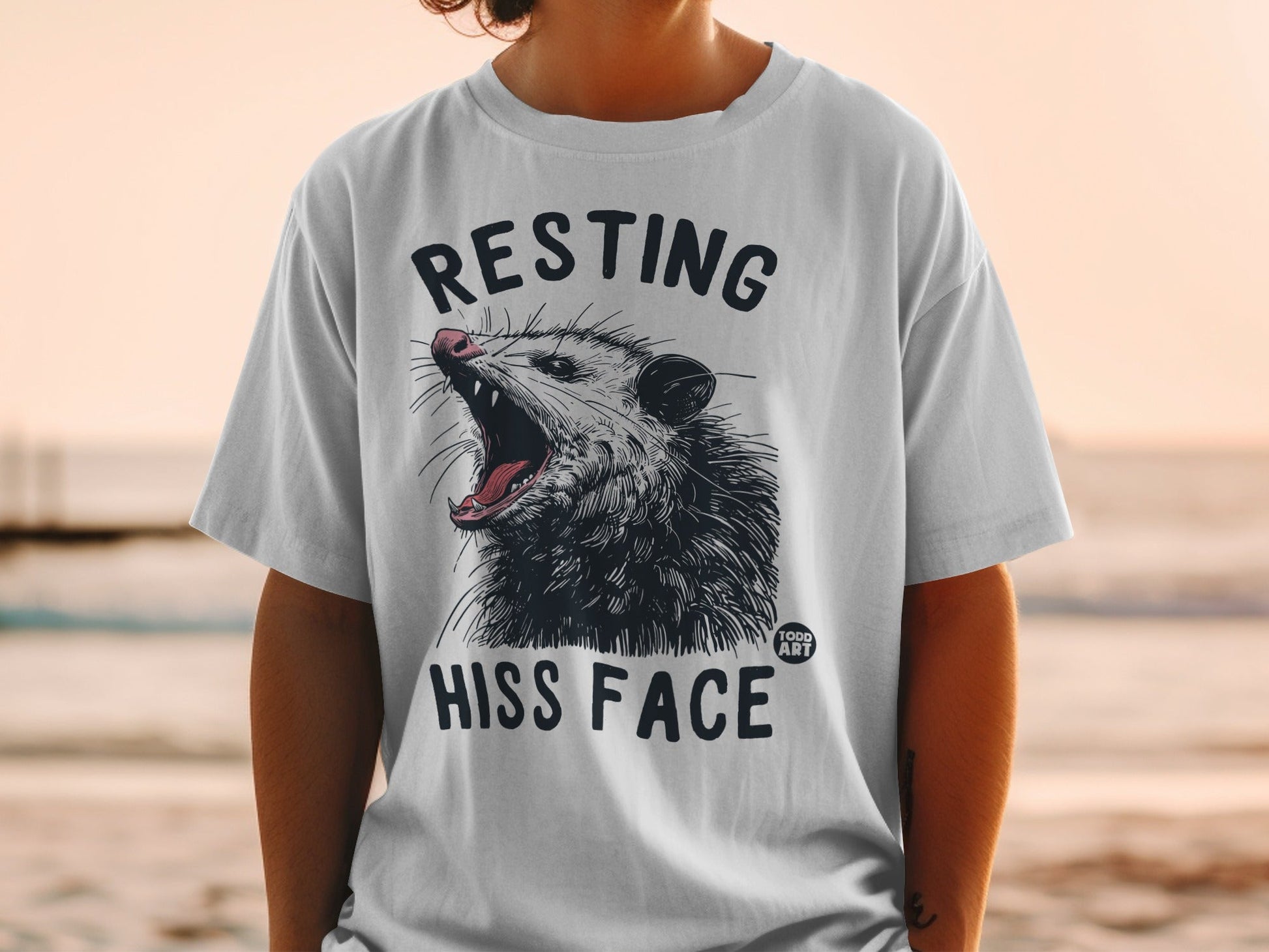 White t-shirt with a graphic of a hissing possum and text "RESTING HISS FACE".