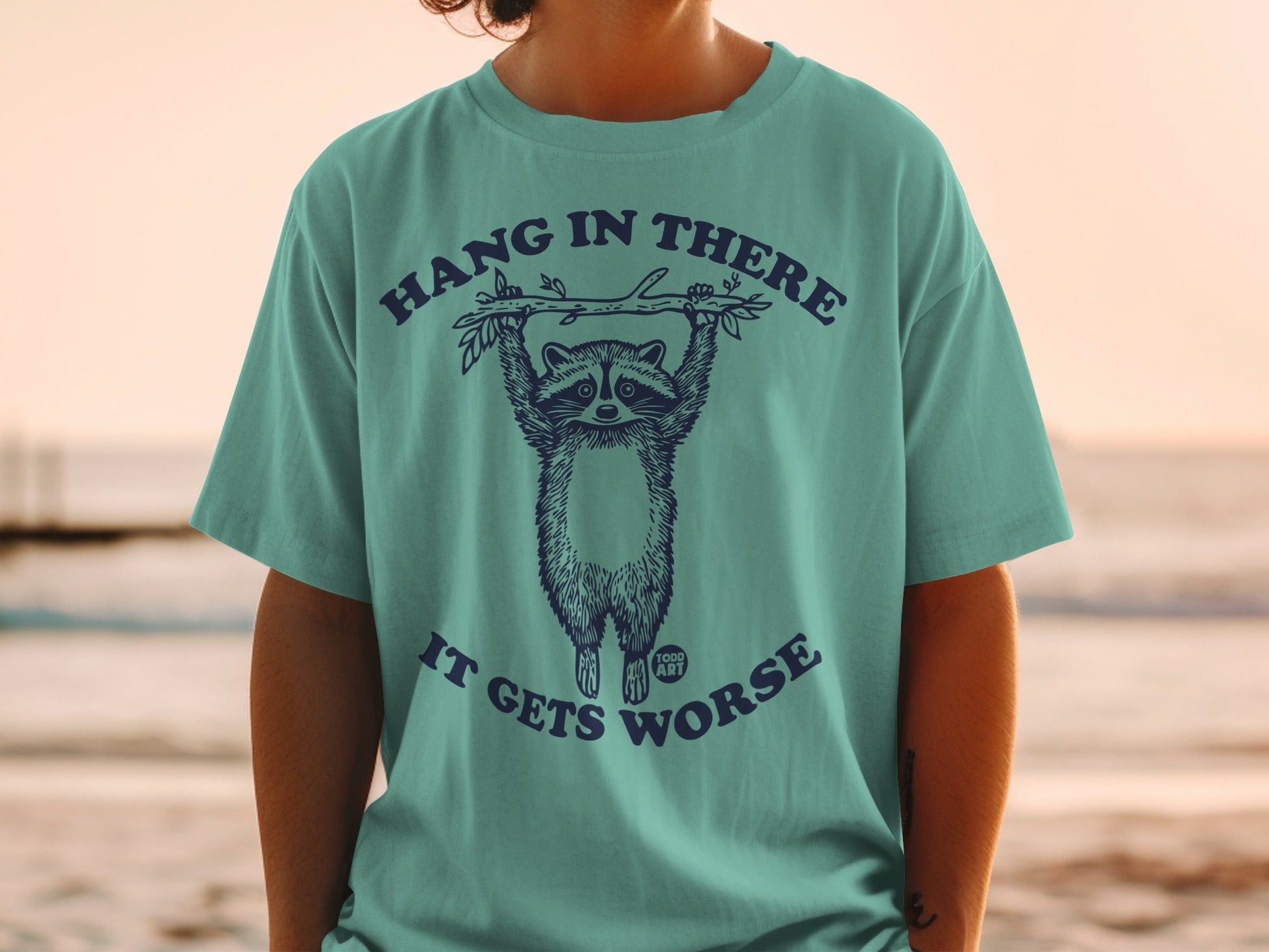 T-shirt with raccoon hanging from a branch saying "Hang in there, it gets worse."