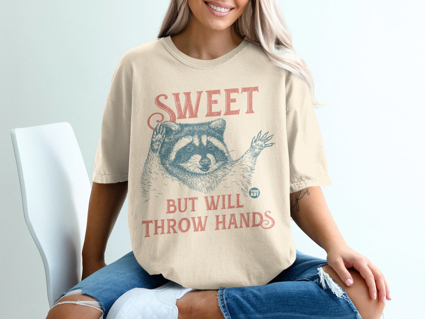 Cream t-shirt with raccoon graphic and text "SWEET BUT WILL THROW HANDS".