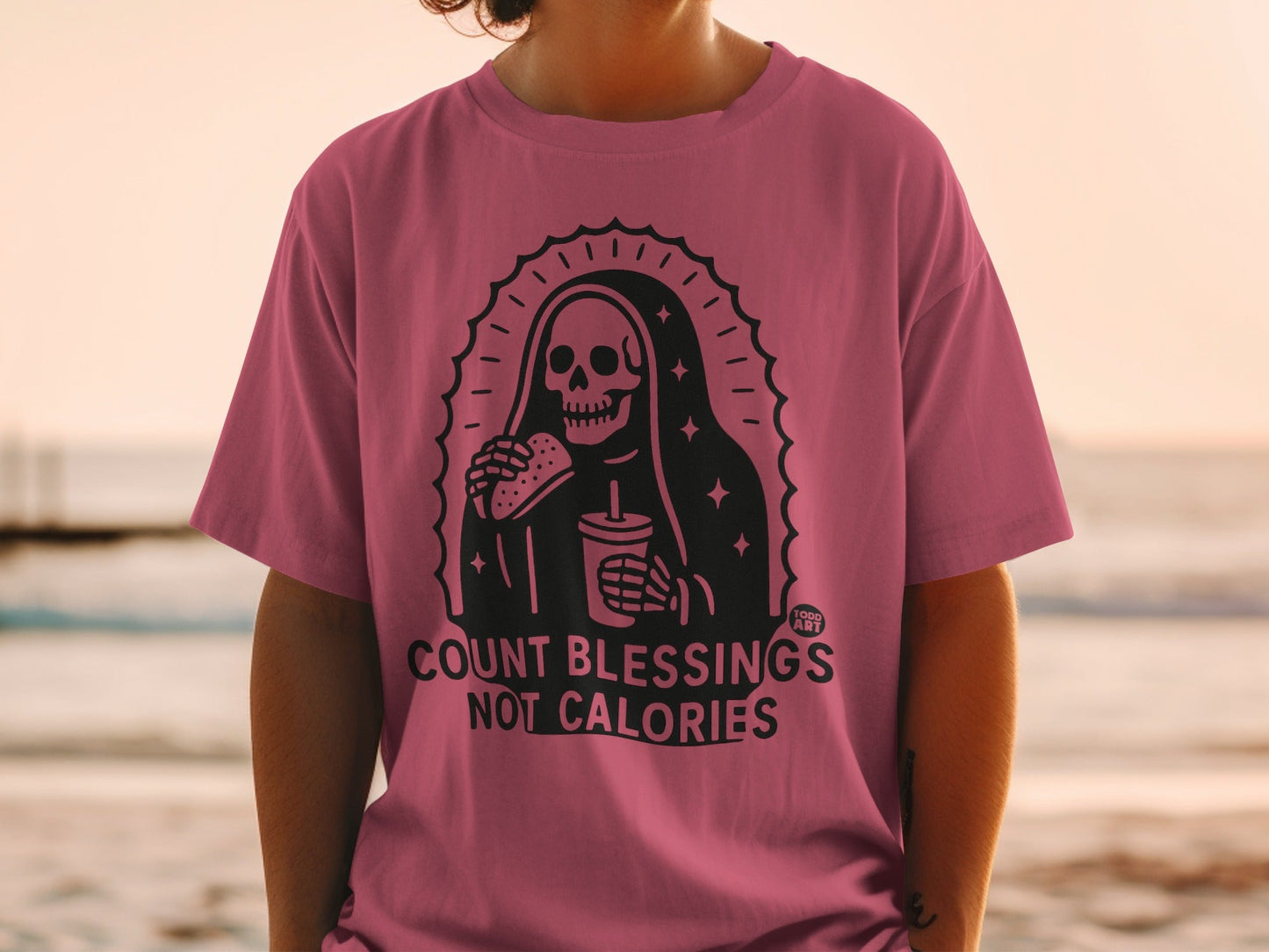 Pink t-shirt with skeleton holding burger and drink, text "COUNT BLESSINGS NOT CALORIES".