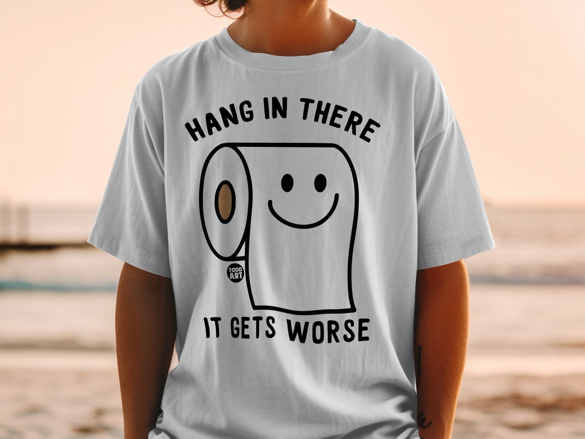 White t-shirt with text "HANG IN THERE IT GETS WORSE" and a smiling toilet paper roll.