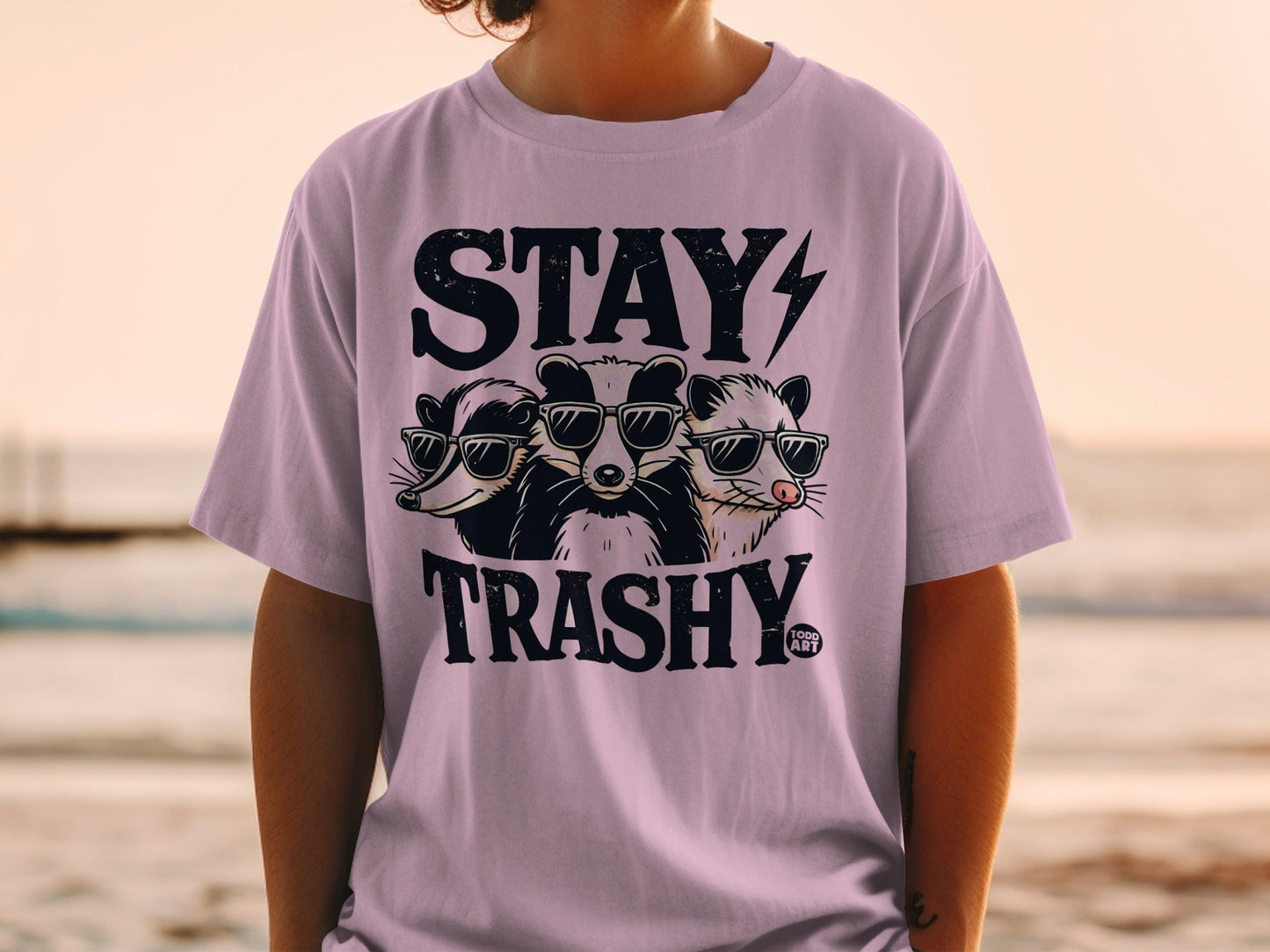 Three raccoons in sunglasses with "STAY TRASHY" text on a purple t-shirt.