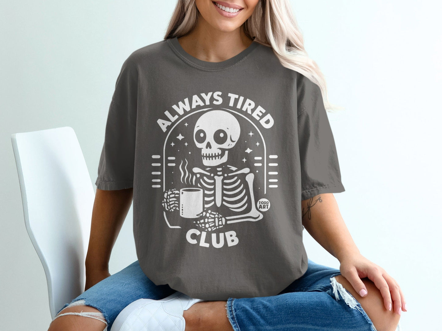 Skeleton with coffee mug and text "Always Tired Club" on gray t-shirt.